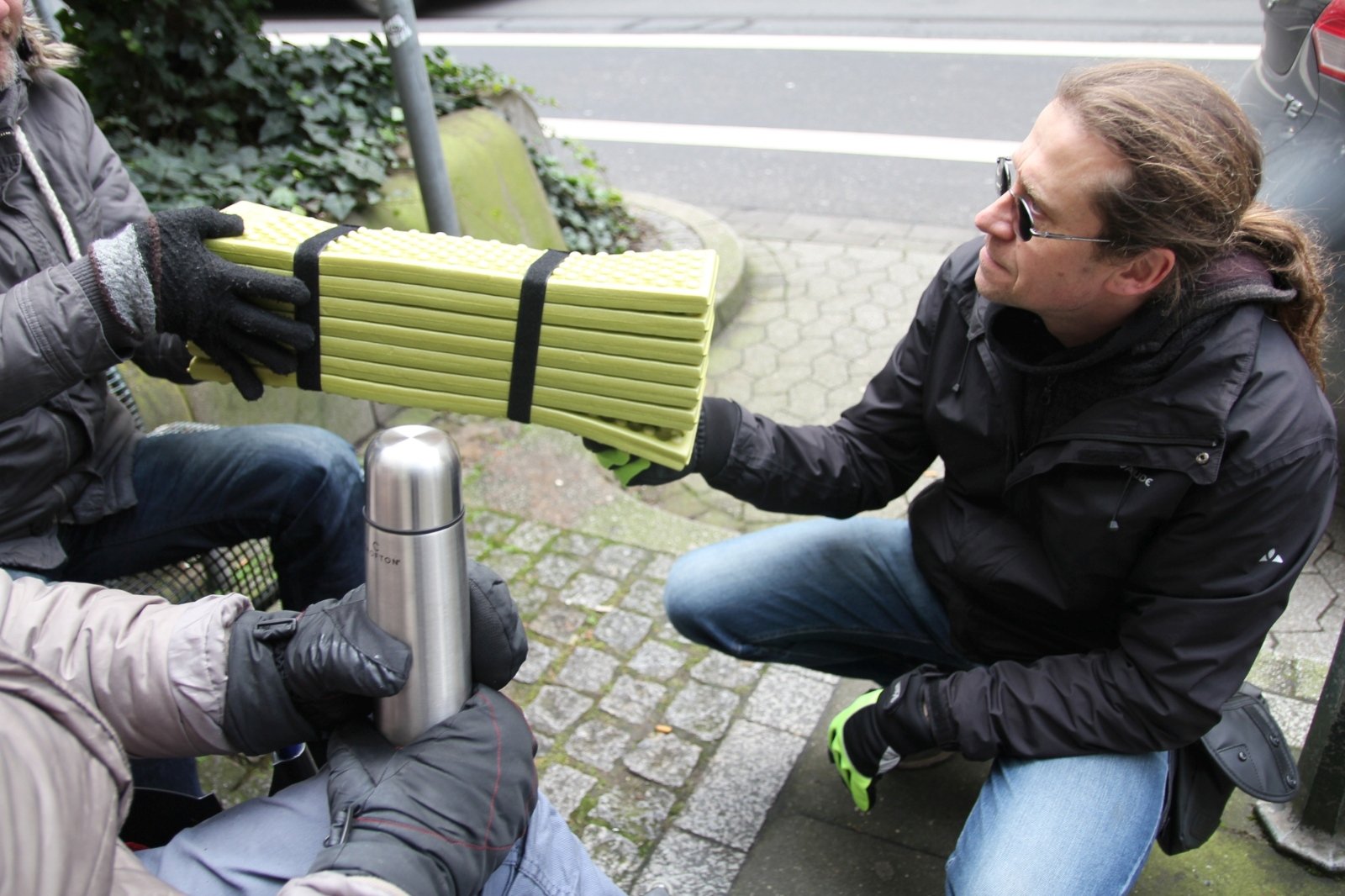 Streetworker Gregor Kozlowski vom Caritasverband Düsseldorf übergibt zwei Obdachlosen Isomatten und eine Thermoskanne mit heißem Tee. (c) CV Düsseldorf